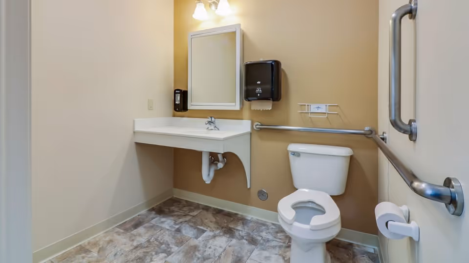 Accessible bathroom with beige walls and tiled floor, featuring a white toilet with grab bars on both sides, a wall-mounted sink with exposed plumbing, a mirror above the sink, a soap dispenser, and a paper towel dispenser.