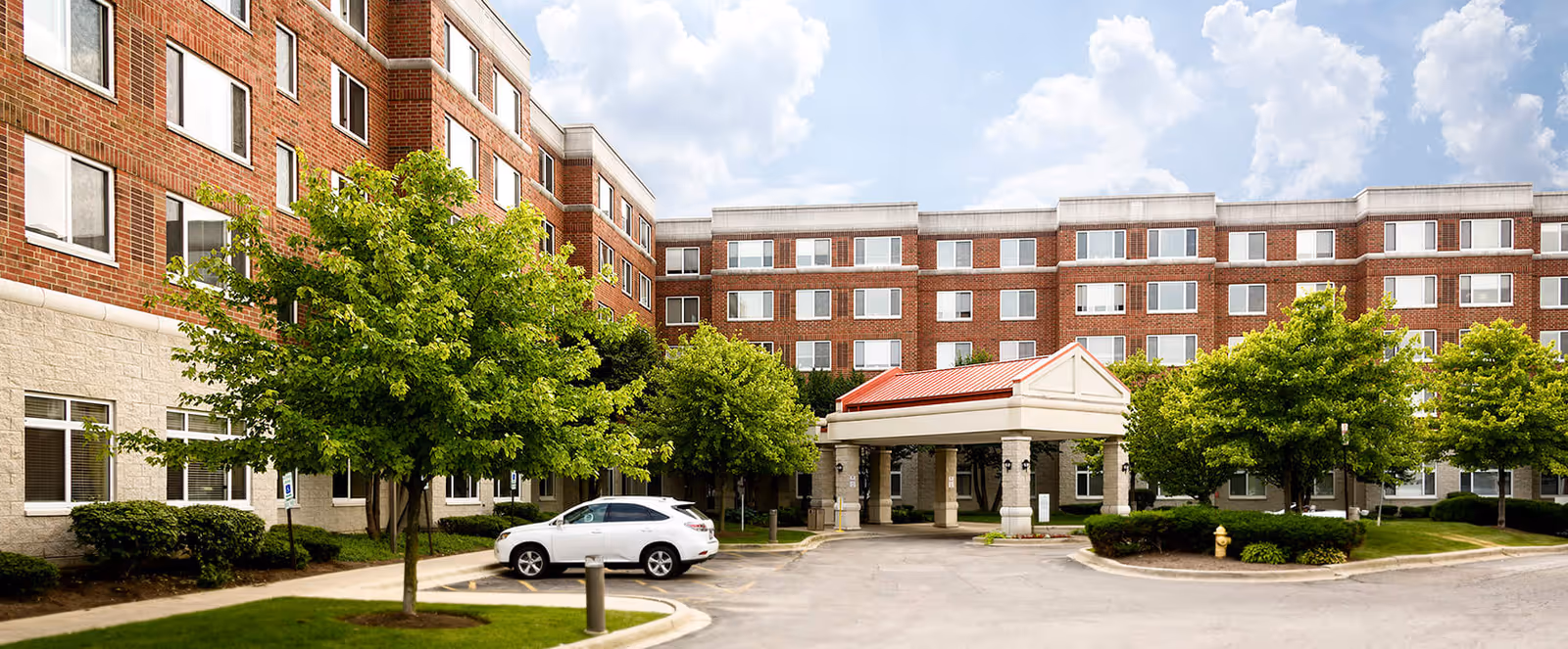 Exterior view of a multi-story senior living facility building with a covered entrance, surrounded by green trees and a parked white car in front.