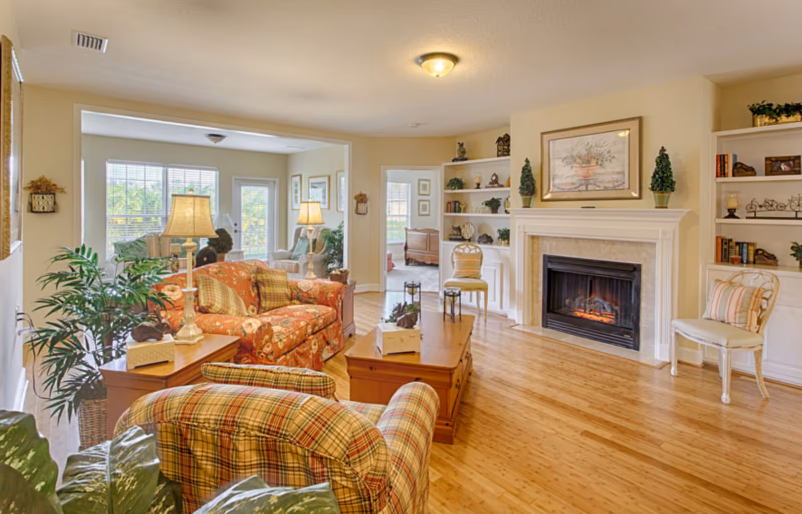 A bright and cozy living room in a senior living facility featuring a floral-patterned sofa, a plaid armchair, wooden coffee and side tables with lamps, a fireplace with a framed painting above it, built-in white shelves decorated with plants and books, and large windows letting in natural light.
