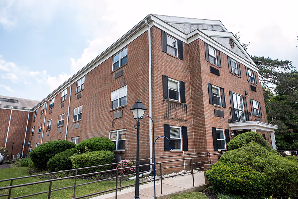 Exterior view of a three-story brick building with multiple windows, black shutters, and a small entrance porch. The building is surrounded by green bushes and a lawn, with a lamppost and a metal handrail along a concrete pathway leading to the entrance. The sky is partly cloudy.