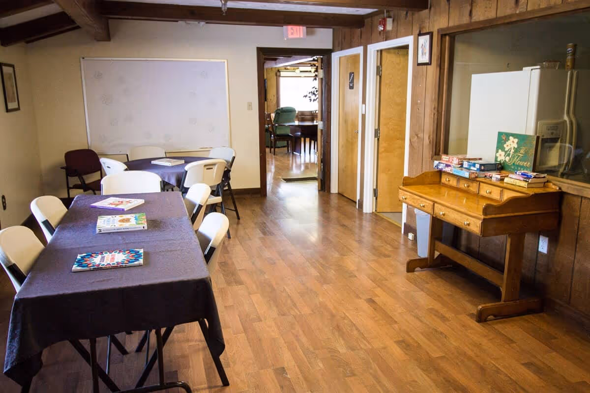 Interior view of a common area with wooden floors and wood-paneled walls. There are two tables covered with dark tablecloths, surrounded by white and maroon chairs. On the tables are several books or magazines. To the right, there is a wooden desk with drawers holding board games and a decorative sign. In the background, there is a hallway leading to another room with more chairs and a table visible.