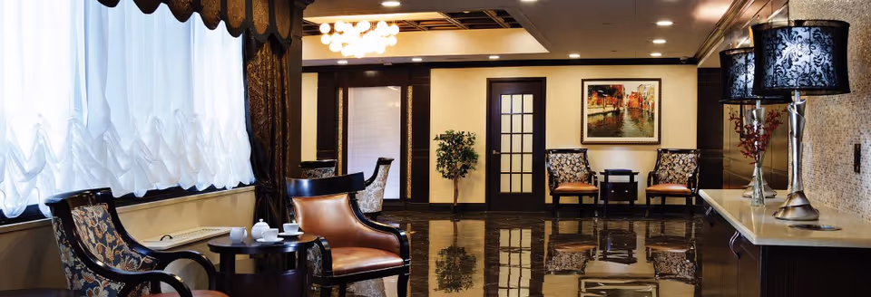 A well-lit, elegant common area with polished dark floors, several upholstered chairs with floral and leather patterns, small round tables with tea sets, a decorative lamp with a patterned shade on a countertop, a potted plant, and framed artwork on the wall.