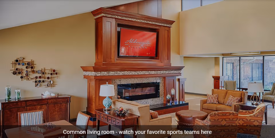 A spacious common living room with comfortable seating including sofas and armchairs arranged around a large wooden fireplace with a mounted TV above it. The room has large windows letting in natural light and decorative wall art on one side. There is a table with chairs and a chessboard in the foreground.