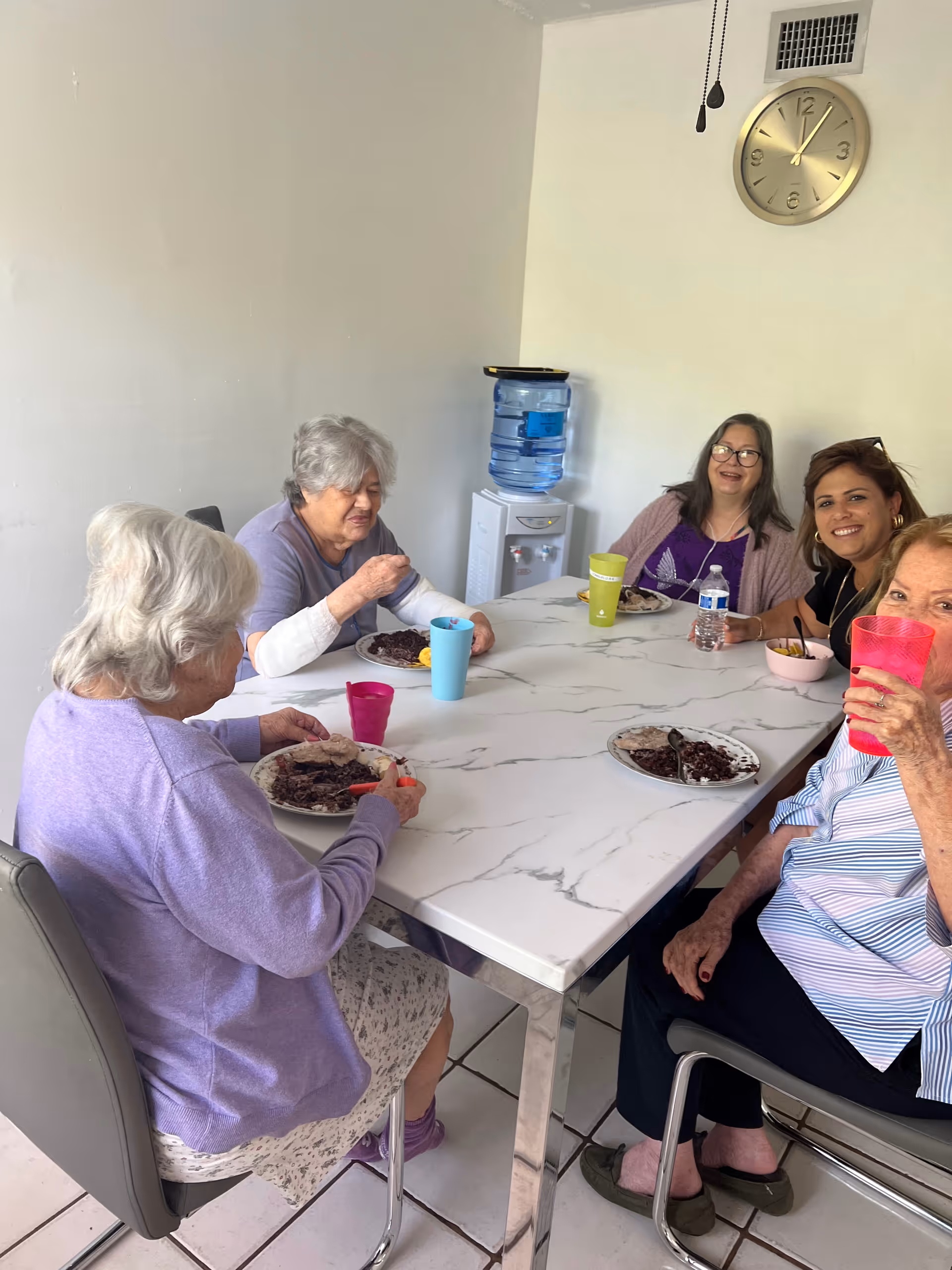 Five elderly women sitting around a rectangular dining table with a marble-like surface, eating a meal together. There is a water dispenser in the corner of the room and a wall clock showing the time as 12:05. The women appear to be enjoying their time, with some smiling and one holding a pink cup.