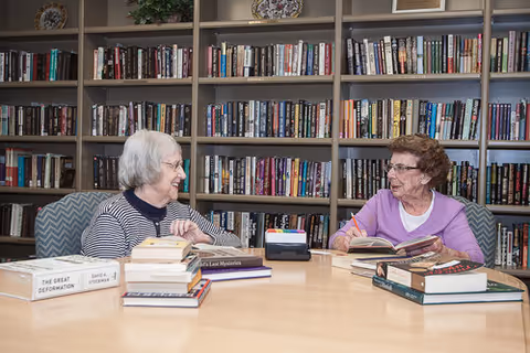 Two elderly women sitting at a round table in a library or reading room, engaged in conversation with books open in front of them. Behind them are shelves filled with numerous books.