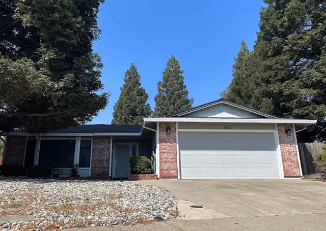 Front exterior view of a single-story house with a two-car garage, brick and siding facade, surrounded by tall trees under a clear blue sky.