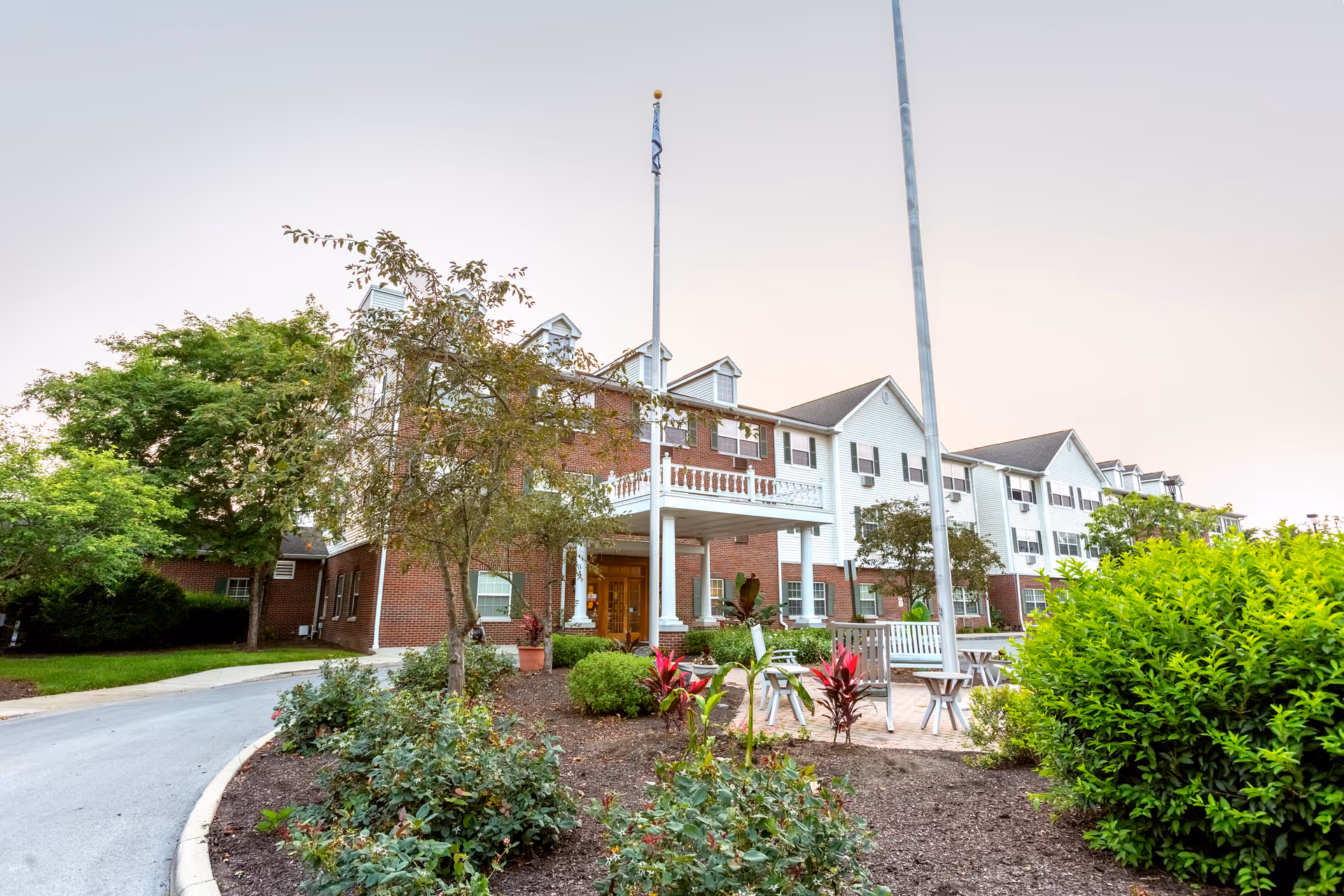 Exterior view of a multi-story senior living facility building with a brick and white facade, surrounded by landscaped greenery, trees, and a paved driveway. There are outdoor seating areas with benches and chairs near the entrance under a covered porch with white columns.