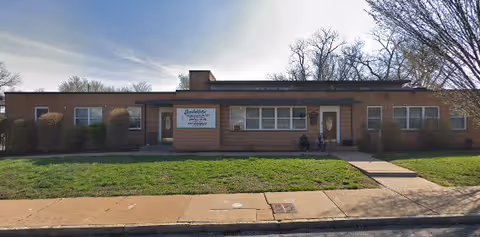 Single-story brick building with a flat roof, identified as Saddler Residential Care by a sign on the front. There is a sidewalk and a grassy area in front of the building, with two people sitting near the entrance. Trees without leaves are visible in the background under a partly cloudy sky.
