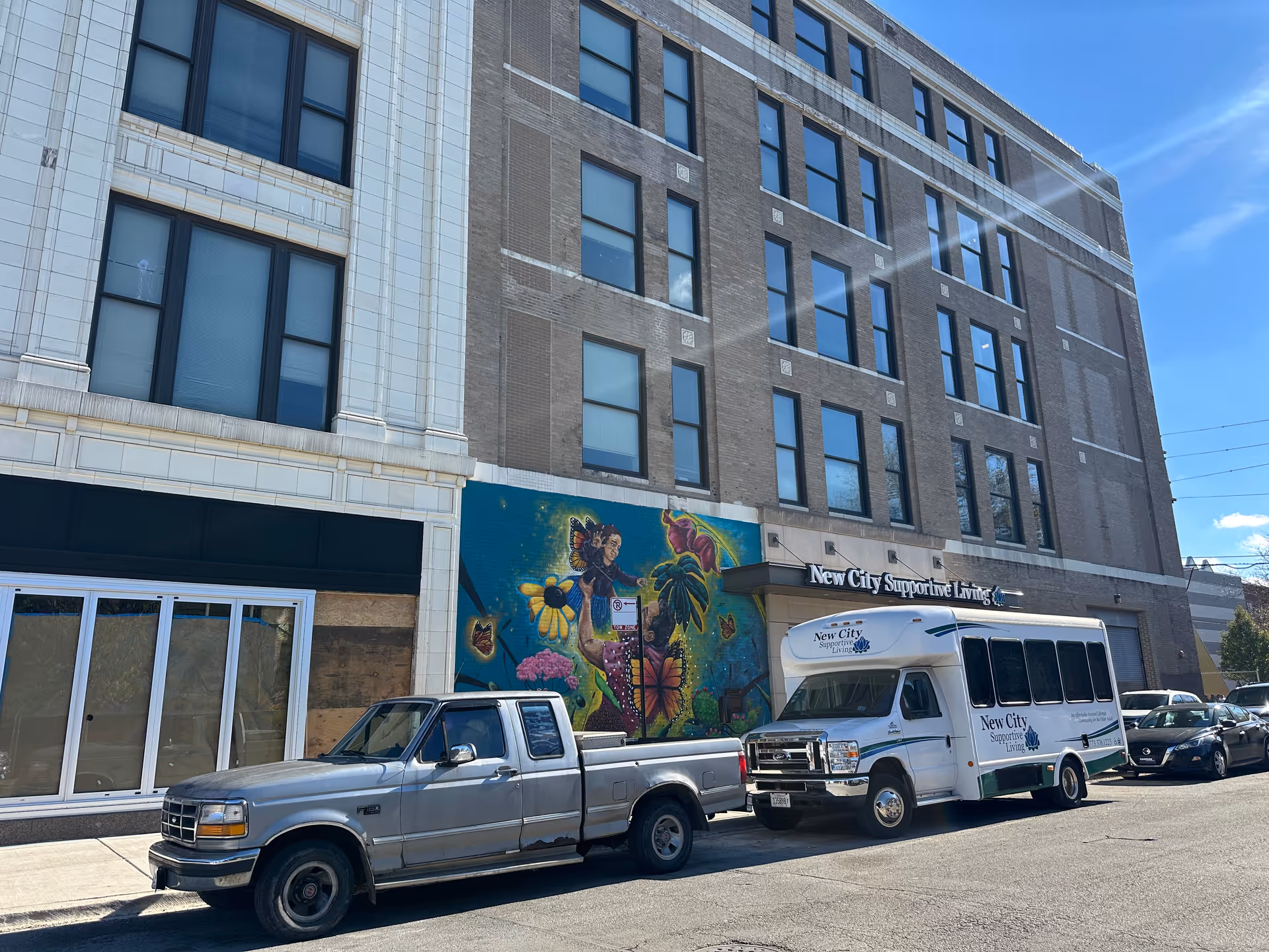 Exterior view of a multi-story brick building with a mural depicting a woman holding a child surrounded by flowers and butterflies. Parked in front are a silver pickup truck and a white shuttle bus with the logo and name 'New City Supportive Living'. The building has a sign with the same name above the entrance.