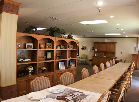 A long dining table with multiple chairs arranged around it in a well-lit room. The room features wooden shelving units with framed photos, decorative items, and plants on top. The walls are light-colored with a patterned wallpaper border near the ceiling. There is a cabinet and additional chairs in the background.