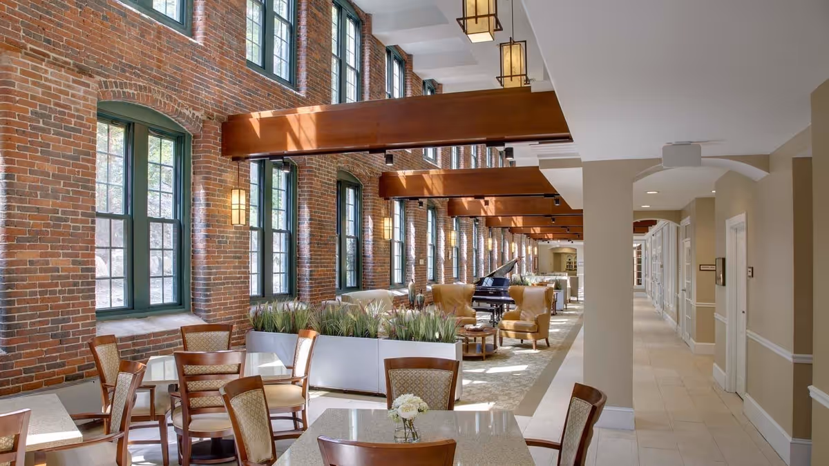 A bright and spacious common area with large windows and exposed brick walls. The room features several tables and chairs in the foreground, a row of planters with green plants, comfortable armchairs, and a grand piano in the background. Wooden beams and hanging light fixtures add architectural interest to the space.