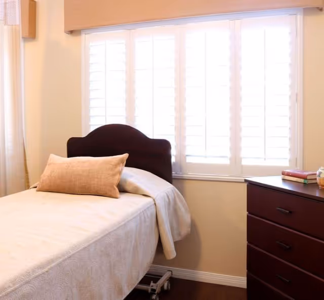 A single bed with a beige bedspread and a matching pillow in a room with a large window covered by white plantation shutters. Next to the bed is a dark wooden dresser with a few books and a small decorative item on top.
