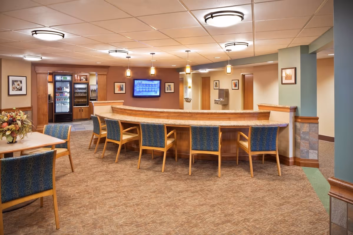 Interior view of a senior living facility lounge area with a curved wooden counter and several blue upholstered chairs. There is a table with chairs and a floral arrangement on the left side. The background shows a small kitchenette area with refrigerators and a wall-mounted TV displaying a schedule. The walls are decorated with framed artwork and the lighting is warm with ceiling fixtures and hanging pendant lights.