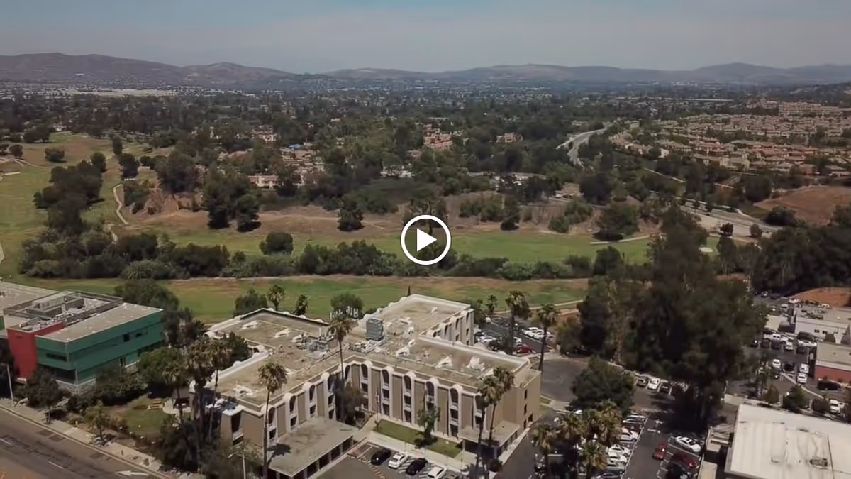 Aerial view of The Pavilion at Sunny Hills facility surrounded by trees, parking lots, and nearby buildings with a backdrop of hills and a sprawling suburban area.