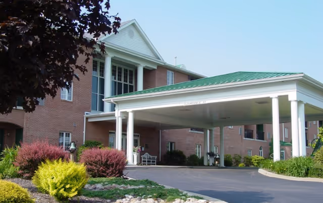 Exterior view of a brick senior living facility with a green metal roof and a covered entrance supported by white columns. There are landscaped bushes and plants in front of the building under a clear blue sky.