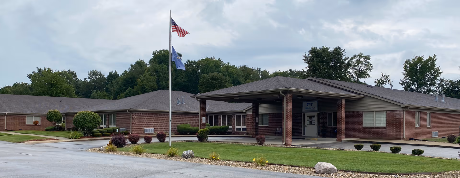 Exterior view of a single-story brick building with a covered entrance and two flagpoles flying the American flag and another flag, surrounded by well-maintained grass, shrubs, and trees under a cloudy sky.