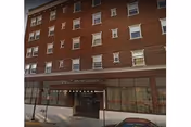 Exterior view of a multi-story brick building with several windows and an entrance canopy labeled Hotel Pennsylvania Apartments. A sidewalk and parked cars are visible in front of the building.
