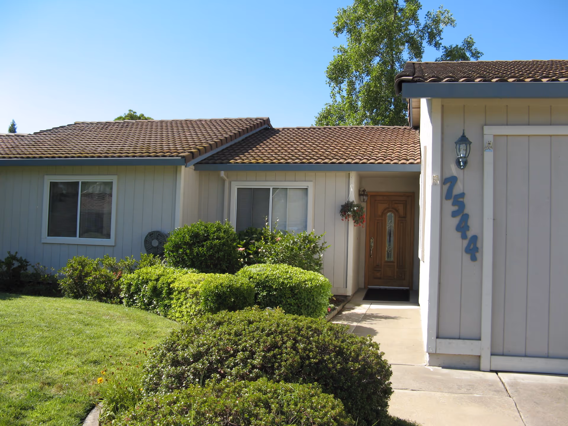 Front exterior view of a single-story house with a tiled roof, beige siding, a wooden front door, and the house number 7544 displayed vertically. There are green bushes and a well-maintained lawn in front of the house under a clear blue sky.