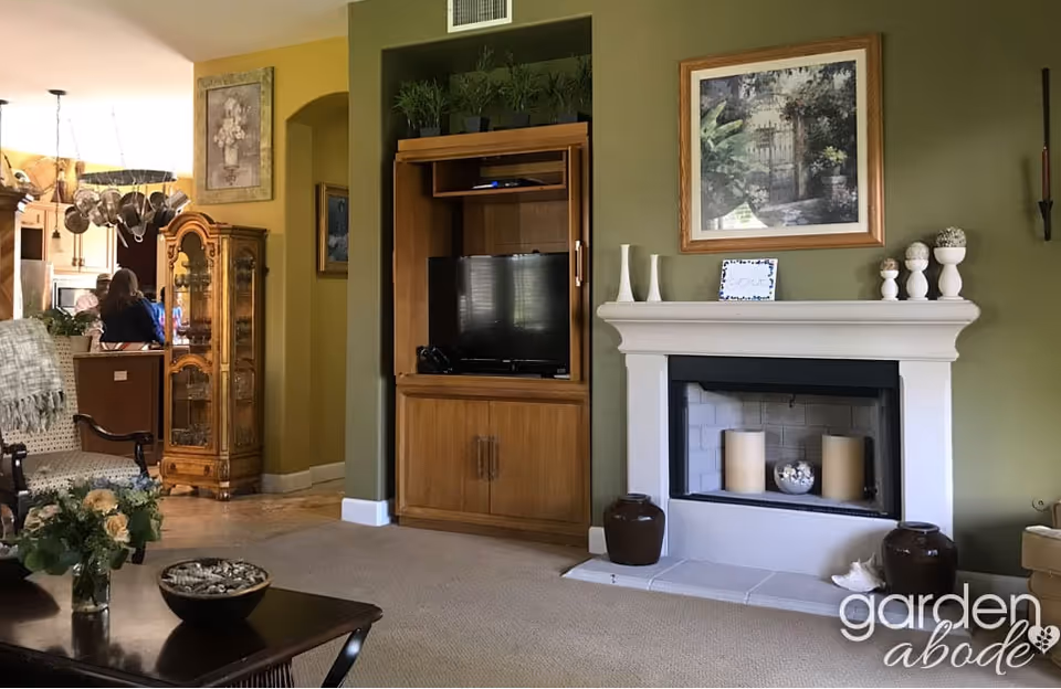 A cozy living room with a green accent wall featuring a white fireplace decorated with candles and vases. Next to the fireplace is a wooden entertainment center with a TV. The room has a beige carpet and a dark wooden coffee table with a flower vase and a bowl. In the background, there is a glimpse of a kitchen area with a hanging pot rack and a person sitting at a counter.