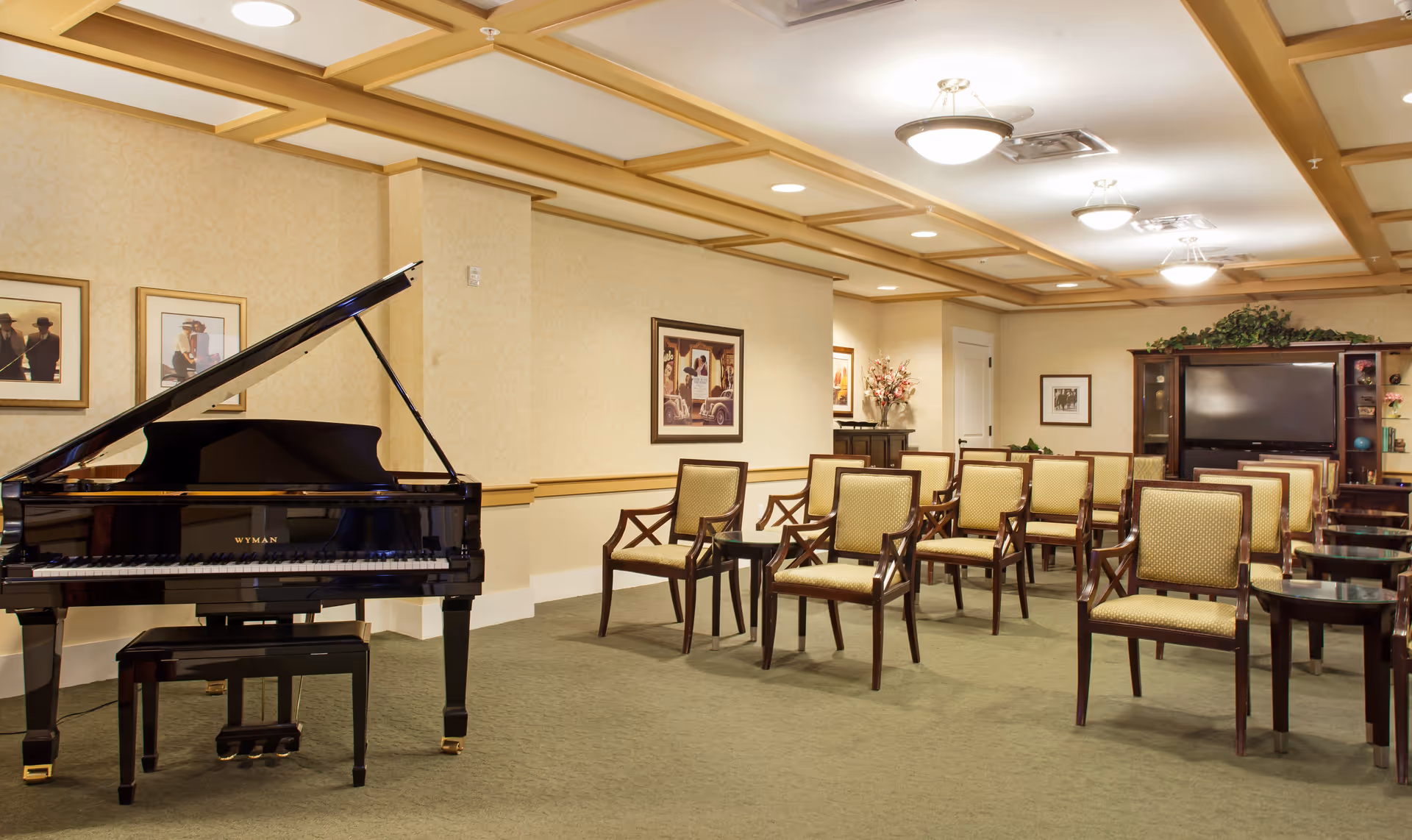 A well-lit room with a black grand piano on the left and several rows of wooden chairs with yellow upholstery arranged facing a large flat-screen TV on a wooden entertainment center. The walls are decorated with framed pictures and floral arrangements, and the ceiling has recessed lighting with wooden beams.