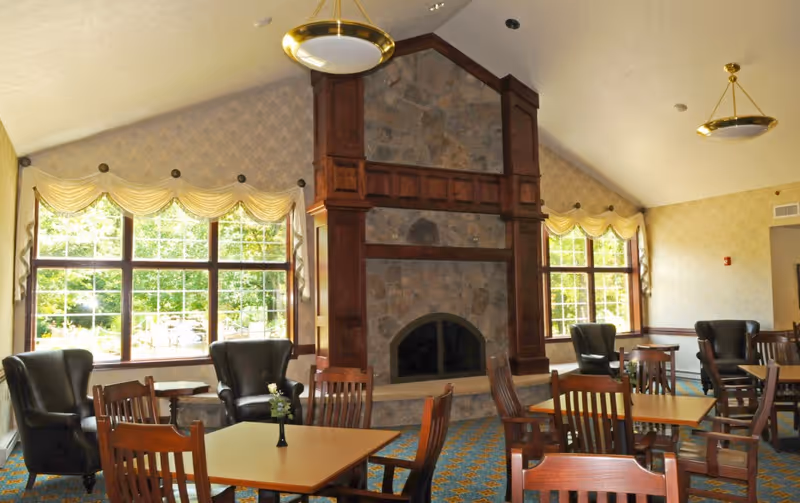 A cozy common area with multiple wooden tables and chairs, several black leather armchairs, large windows with cream-colored valances letting in natural light, and a large stone fireplace with wooden trim as the focal point.