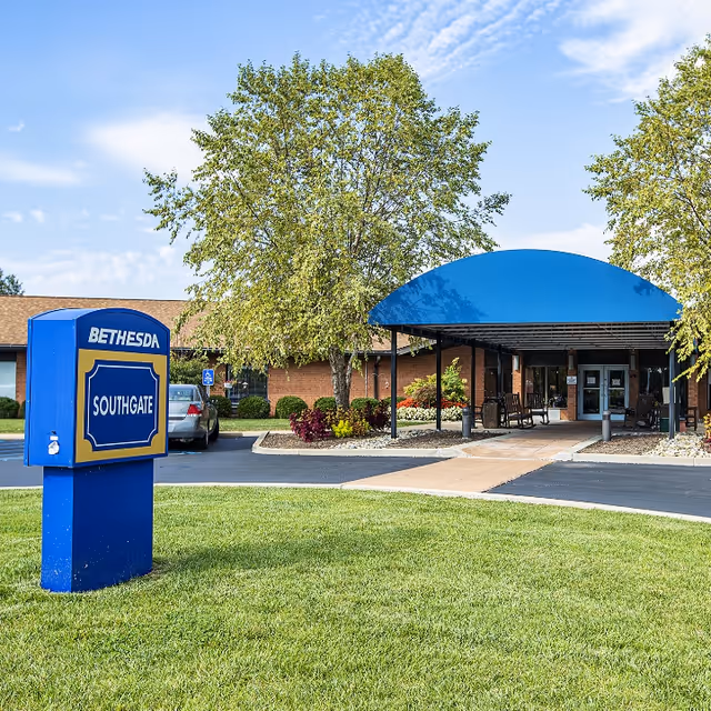 Exterior view of Bethesda Southgate facility showing a blue and yellow sign with the facility name on a grassy area. The building has a covered entrance with a blue awning, surrounded by trees and landscaping under a partly cloudy sky.
