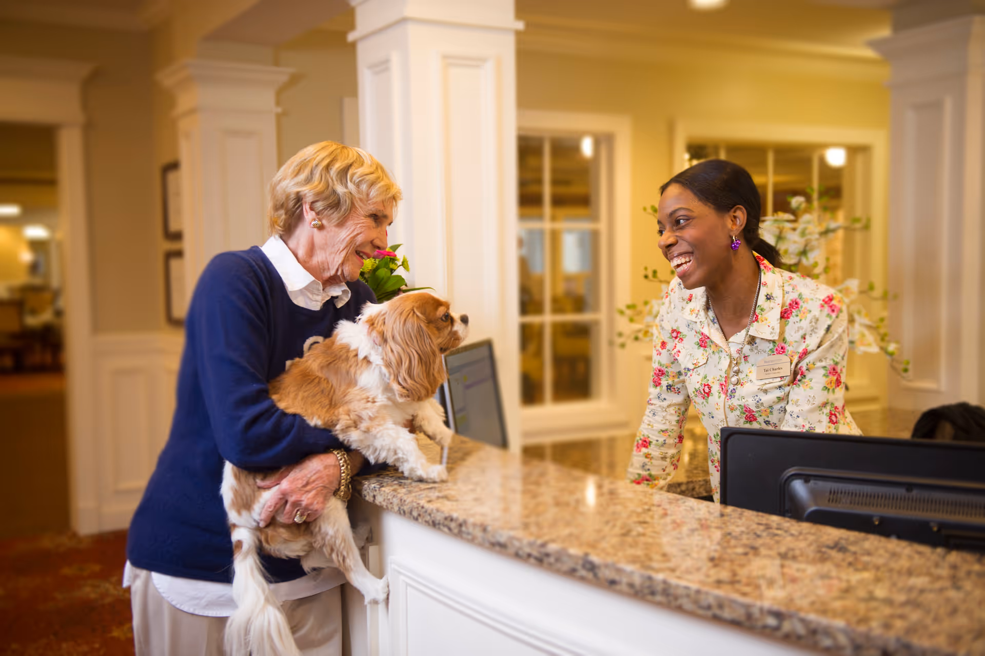 An elderly woman holding a small dog is smiling and talking to a smiling receptionist standing behind a granite countertop in a warmly lit senior living facility lobby.