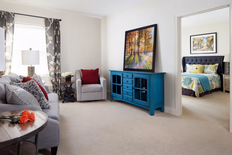 A bright living room with a gray sofa adorned with patterned and red pillows, a patterned armchair with a red pillow, a wooden side table with a flower arrangement, and a blue cabinet with a framed painting of a sunlit forest path resting on top. Through an open doorway, a bedroom is visible with a bed featuring floral bedding, a black tufted headboard, and a framed landscape painting above the bed.