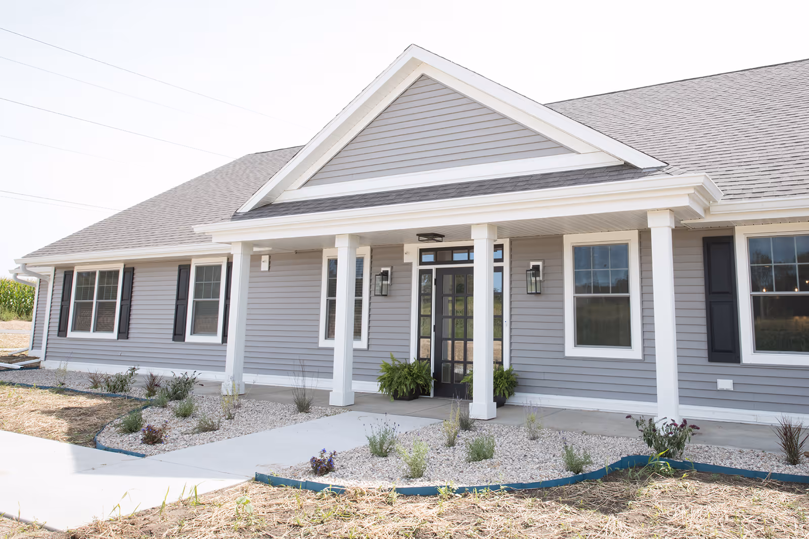 Front exterior view of a single-story building with gray siding, white trim, black shutters, and a covered entrance supported by white columns. There are small landscaped areas with plants and gravel along the walkway leading to the entrance.