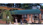 Entrance of a brick building with a green awning covering the walkway. Two people are near the entrance, one standing and one in a wheelchair. There are plants and trees around the entrance area.