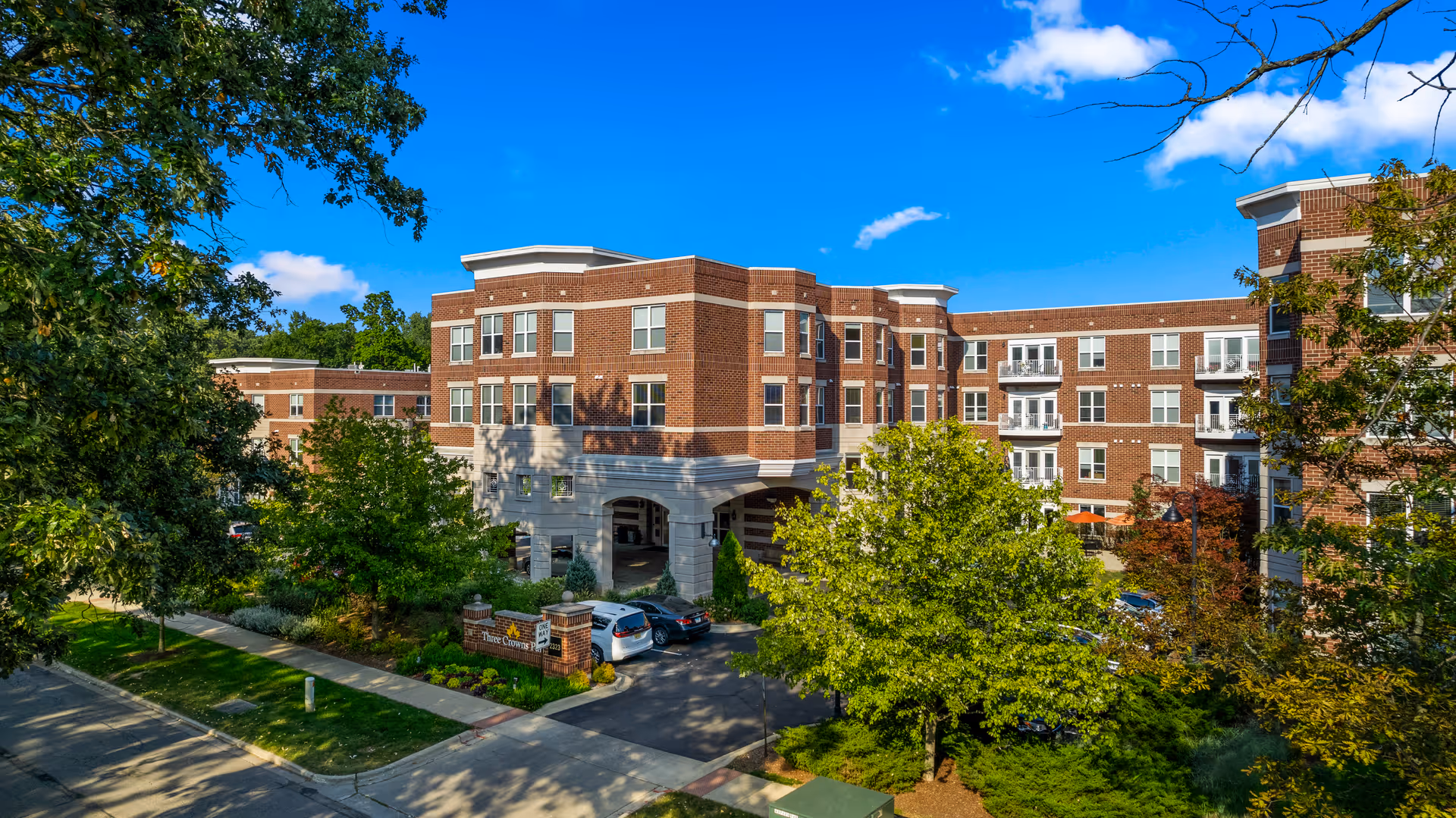 Exterior view of a multi-story brick senior living facility named Three Crowns Park, surrounded by trees and greenery under a bright blue sky with some clouds. Several cars are parked near the entrance.