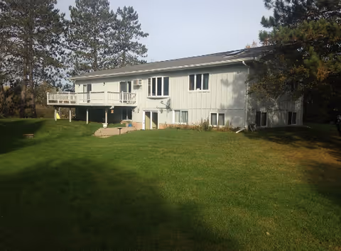 A single-story light gray building with multiple windows and a raised deck on one side, surrounded by a large green lawn and tall trees under a clear sky.