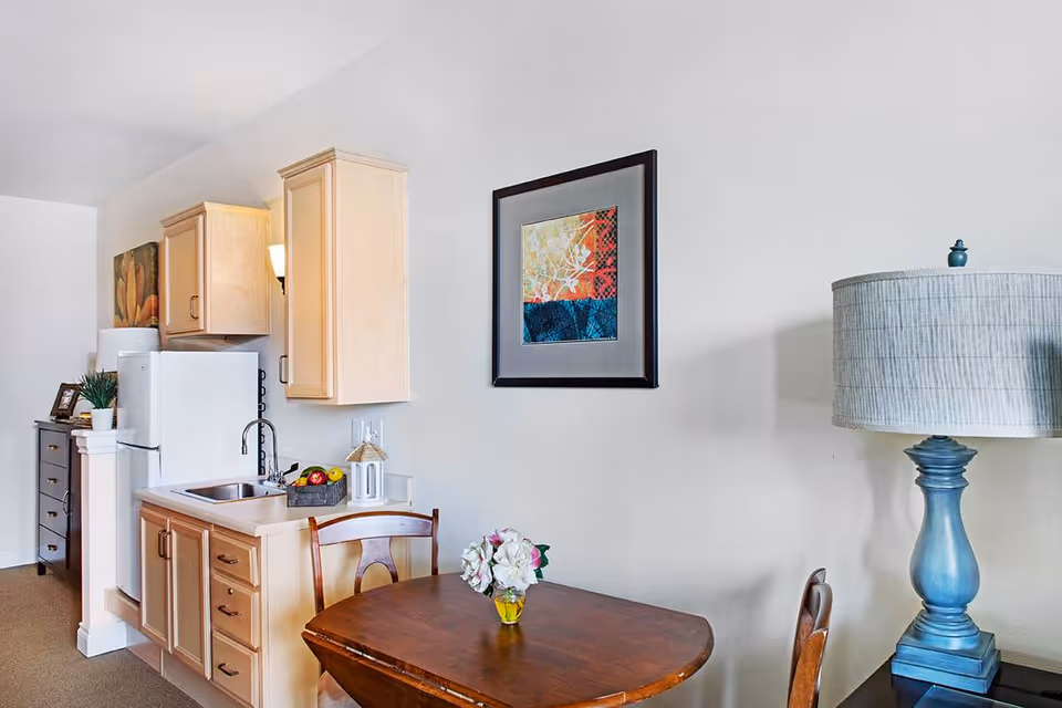 Small kitchen area with light wood cabinets, a white refrigerator, and a sink. A wooden dining table with two chairs is in front, with a small vase of flowers on the table. A blue lamp is on a side table to the right, and a framed colorful abstract artwork hangs on the wall above the dining table.