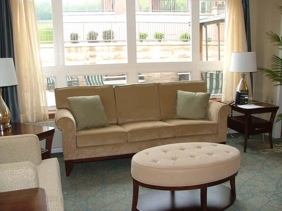 A cozy living room area with a beige sofa featuring two green cushions, a matching beige oval ottoman, a beige armchair, two wooden side tables each with a lamp, and large windows showing an outdoor patio with chairs and tables.