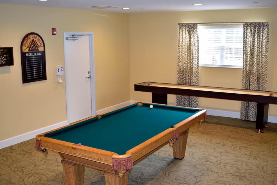 A game room with a pool table in the foreground and a shuffleboard table against the far wall. The walls are painted light yellow, and there is a window with patterned curtains letting in natural light. On the left wall, there is a scoreboard for billiards and darts and a hand sanitizer dispenser next to a closed white door.
