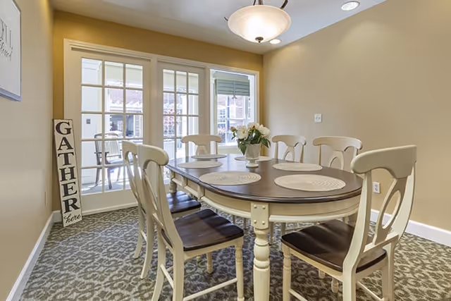 A dining room with a wooden table and six chairs. The table has round placemats and a vase with flowers in the center. There is a sign leaning against the wall that says 'GATHER here'. French doors with glass panes lead to an outdoor area.