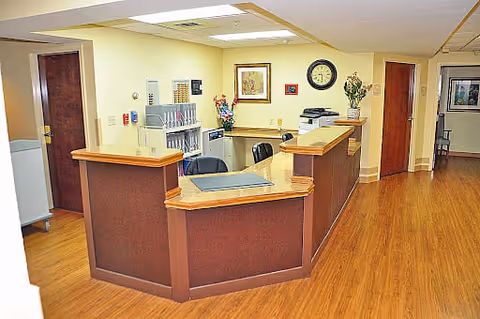 Reception desk area inside a senior living facility with wooden flooring, beige walls, and overhead fluorescent lighting. The desk has a dark wood finish with a lighter wood countertop. Behind the desk are office chairs, a clock on the wall, framed pictures, and some office equipment including a printer. There are doors and a hallway visible in the background.