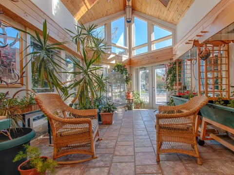 A bright sunroom with large windows and a high wooden ceiling featuring exposed beams. The room has tiled flooring and contains two wicker rocking chairs facing each other, surrounded by various potted plants. There is a glass door at the far end leading outside, allowing natural light to fill the space.