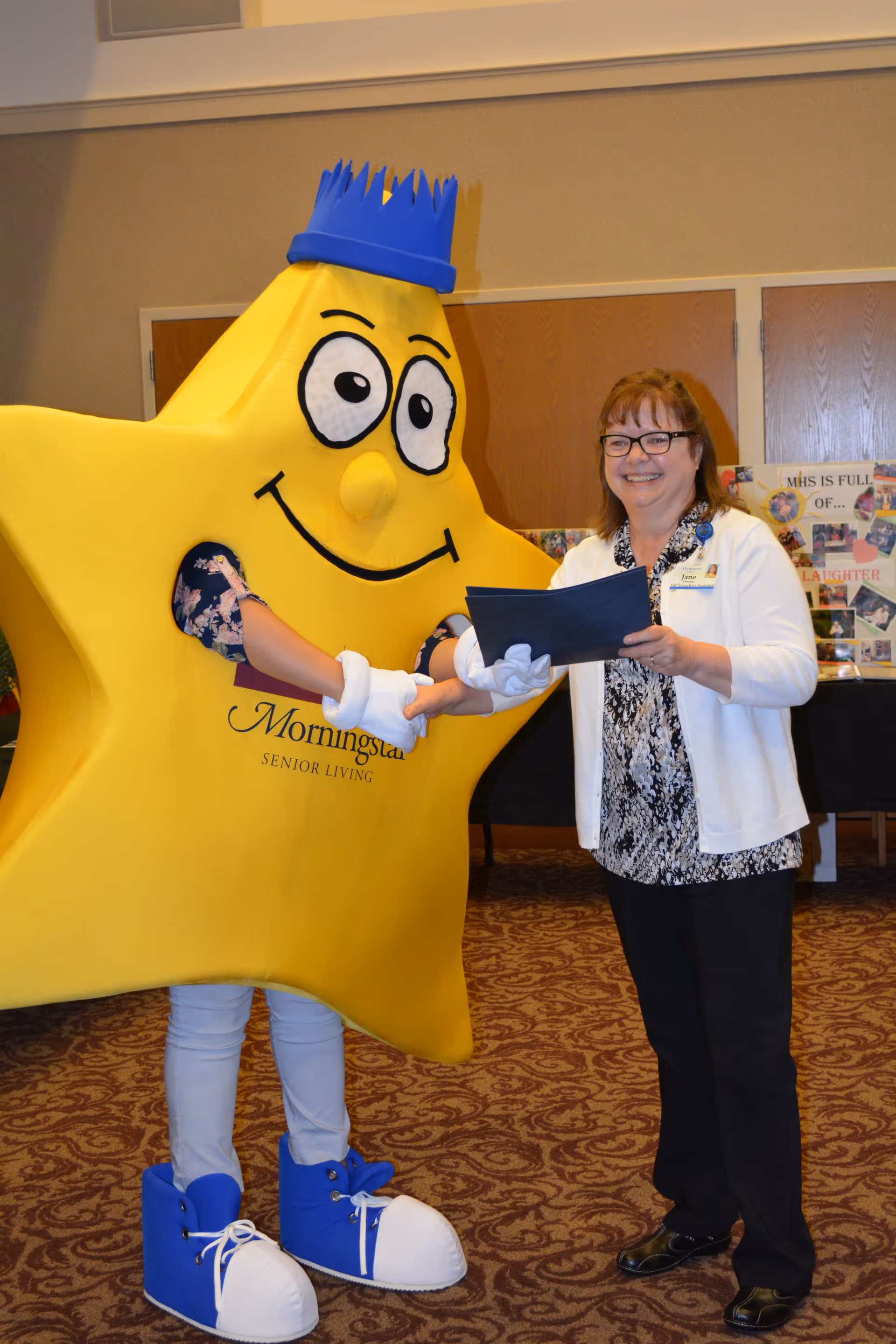 A person dressed in a large yellow star costume with a blue crown and blue shoes shakes hands with a smiling woman wearing glasses, a white cardigan, and black pants inside a room with carpeted floor and wooden doors in the background.
