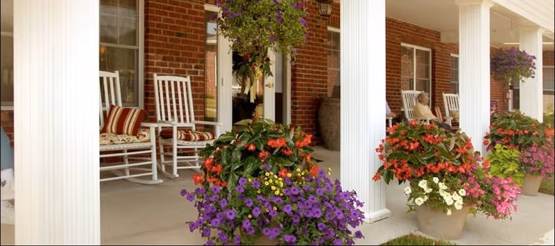 A covered porch area with white columns and several white rocking chairs with striped cushions. Large flower pots with vibrant red, purple, yellow, and white flowers are placed along the porch. A person is sitting on one of the rocking chairs in the background near a brick wall and windows.