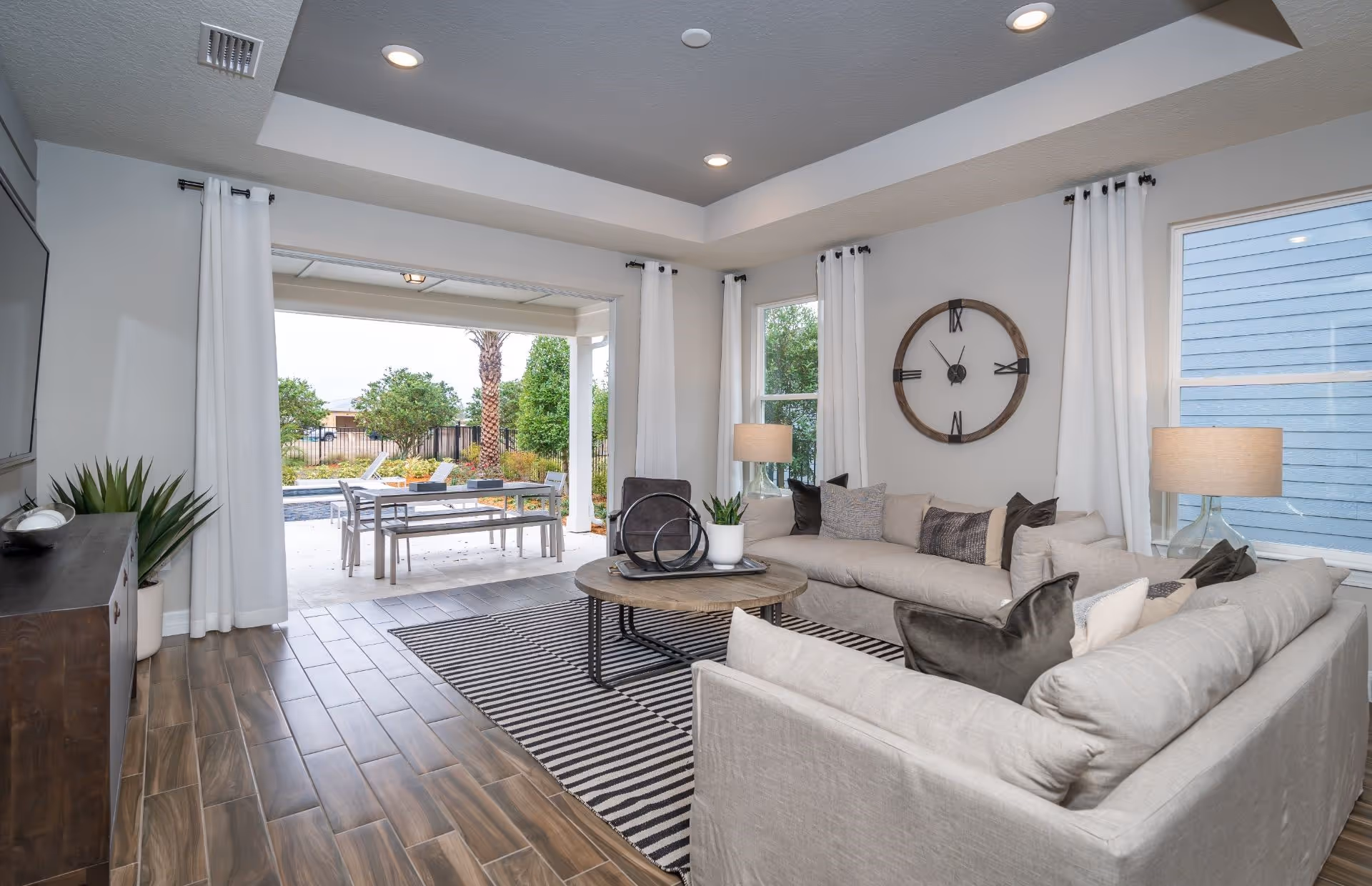 A modern living room with two beige sofas arranged around a round wooden coffee table on a striped rug. The room has wood-look tile flooring, white walls, and a large wall clock. Two windows with white curtains and two table lamps provide lighting. The living room opens to an outdoor patio area with a dining table and chairs, surrounded by greenery and palm trees.