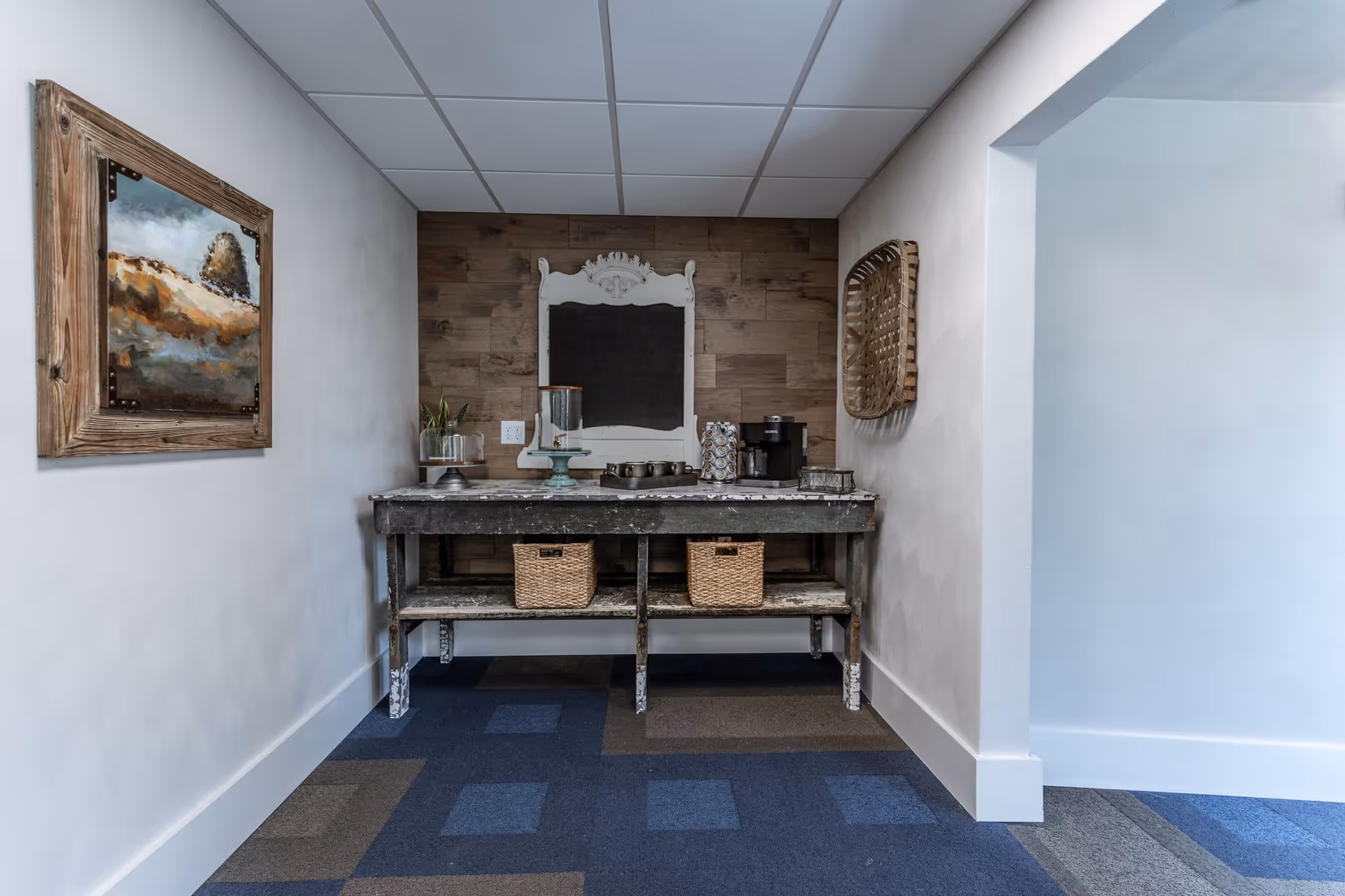A small coffee station area with a rustic wooden table holding a coffee maker, cups, and a glass container. Above the table is a decorative white-framed chalkboard on a wood-paneled wall. To the left, a framed landscape painting hangs on a white wall, and to the right, a woven basket decoration is mounted on the wall. The floor is carpeted with a blue and gray checkered pattern.