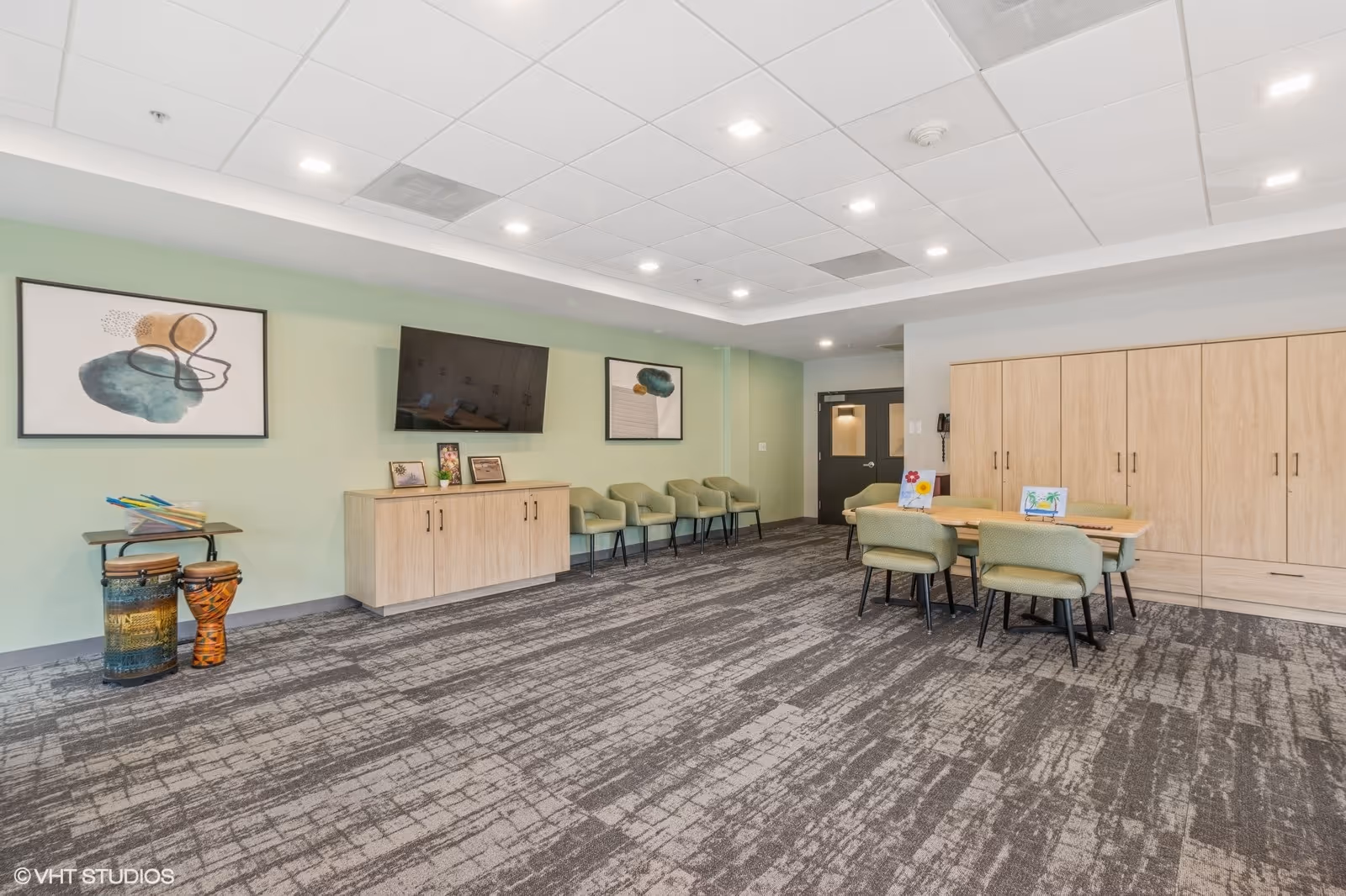 A spacious, well-lit common area with light green walls and patterned gray carpet. The room features a wall-mounted flat-screen TV above a light wood cabinet, four green chairs lined up against the wall, and a table with four green chairs around it. There are two abstract paintings on the wall and two decorative drums near the cabinet. A large wooden storage unit is on the right side of the room.