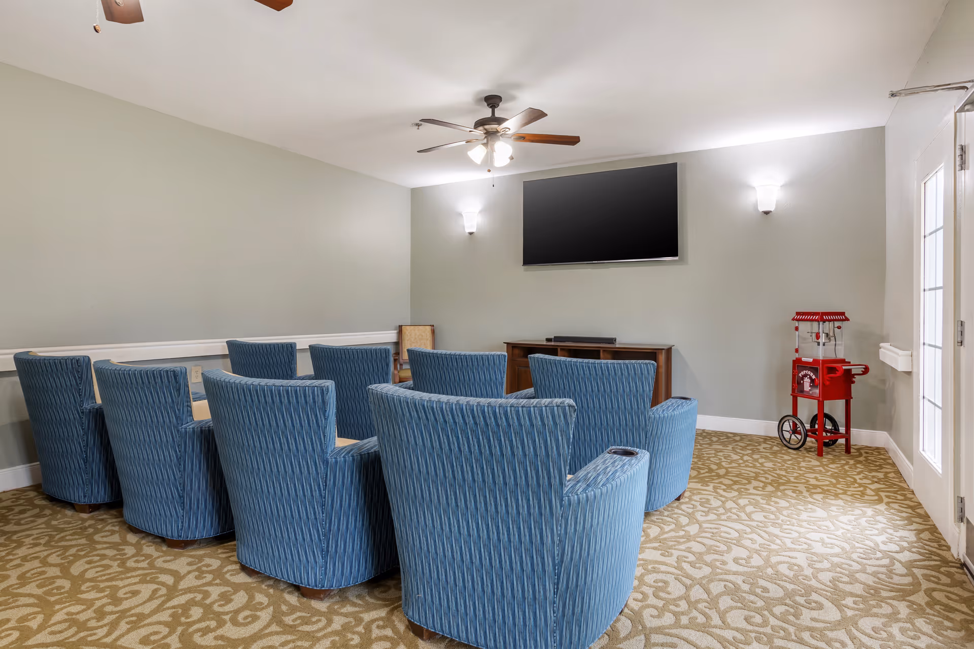 A small theater room with two rows of blue upholstered chairs facing a wall-mounted flat screen TV. The room has beige patterned carpet, light gray walls, a ceiling fan with lights, and a red popcorn machine in the corner near a glass door.