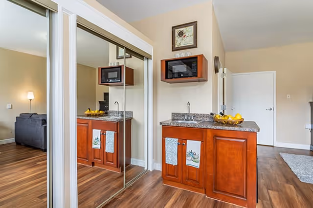 Interior view of a senior living facility room featuring a wooden cabinet with a granite countertop, a sink, and a microwave mounted above. Two towels hang on the cabinet doors. A mirrored sliding closet door reflects the cabinet and part of a living area with a gray sofa and a floor lamp. The room has wood flooring and beige walls.