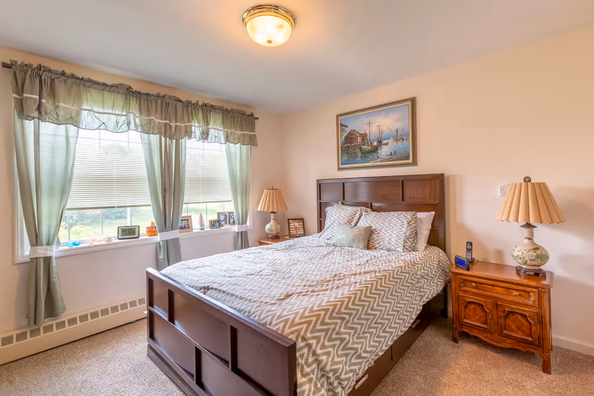 A bedroom with a wooden bed frame and a bed made with a chevron-patterned comforter and multiple pillows. There are two wooden nightstands on either side of the bed, each with a lamp. A large window with green curtains and blinds lets in natural light. A framed painting of boats on water hangs above the bed.