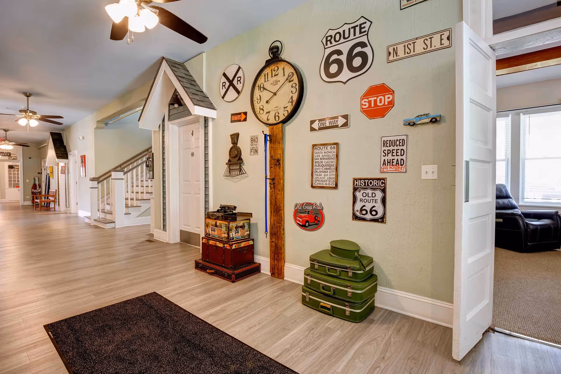 Interior hallway of Shepherd Premier Senior Living with light wood flooring, ceiling fans with lights, and decorative wall featuring vintage Route 66 and street signs, a large clock, and stacked vintage suitcases. There are white doors with small peaked roofs above them and a glimpse into a room with a black recliner chair.