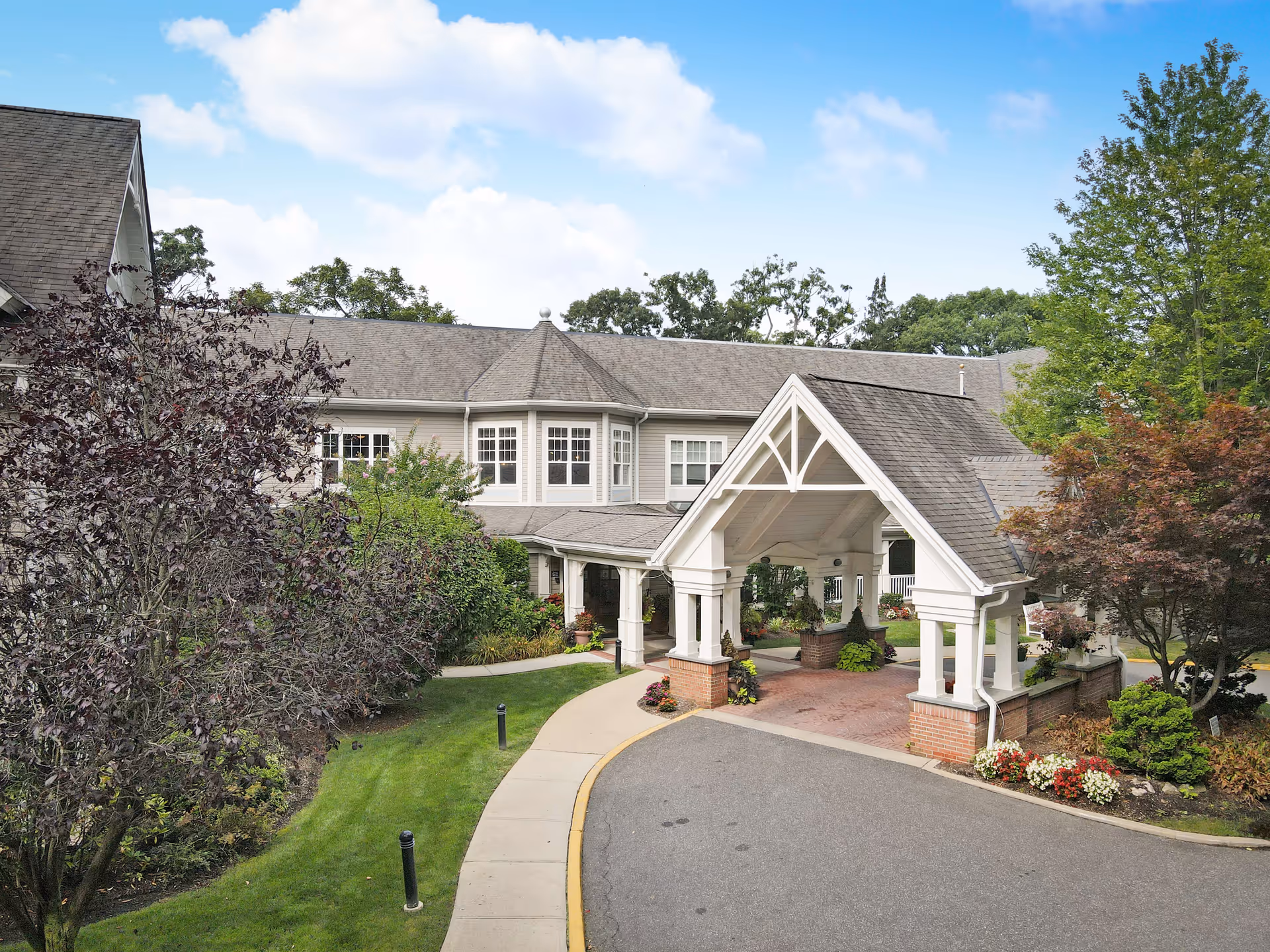 Front exterior view of a senior living facility named Sunrise of Plainview, featuring a covered entrance with white pillars and a peaked roof. The building is surrounded by well-maintained landscaping including trees, shrubs, and flower beds. A curved driveway leads up to the entrance under a partly cloudy sky.