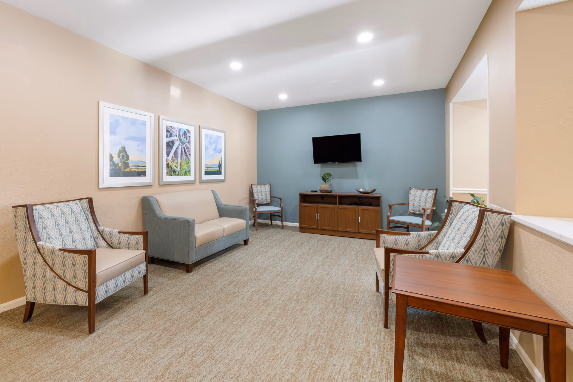 A cozy living room area with beige carpet and walls painted in beige and blue. The room features a small sofa, four armchairs with patterned upholstery, a wooden TV stand with a flat-screen TV mounted on the blue accent wall, and a wooden side table. Three framed landscape paintings hang on the beige wall above the sofa. The room is well-lit with recessed ceiling lights.