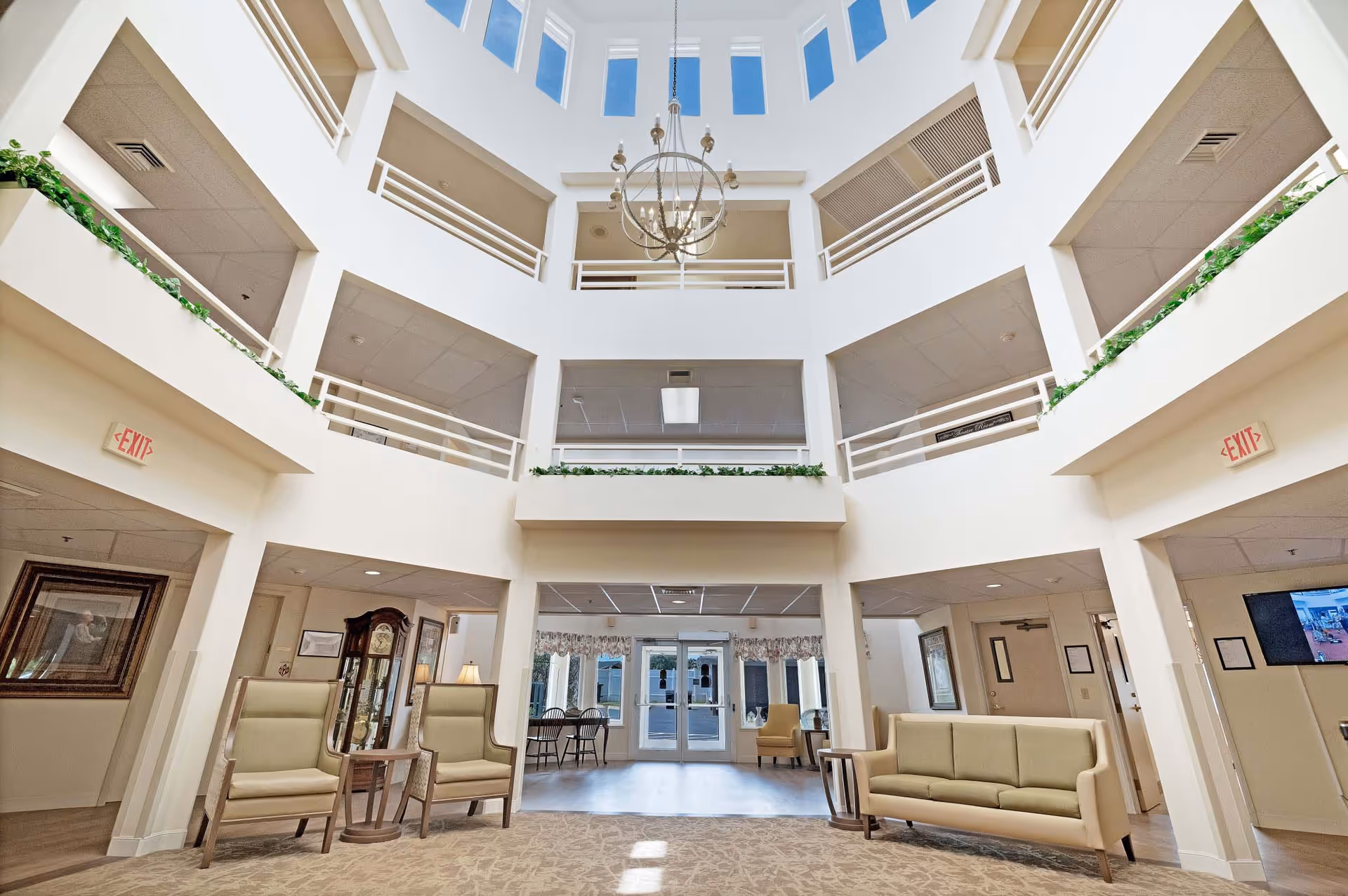 Spacious and bright lobby area of a senior living facility with high ceilings and multiple floors visible. The space includes comfortable seating with two armchairs and a sofa, small side tables, decorative plants along the railings, a chandelier hanging from the ceiling, and large windows letting in natural light. The entrance doors are visible in the background.